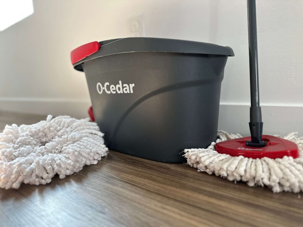 Image of an ocedar mop bucket and ocedar mop against a white wall and brown wooden floors.