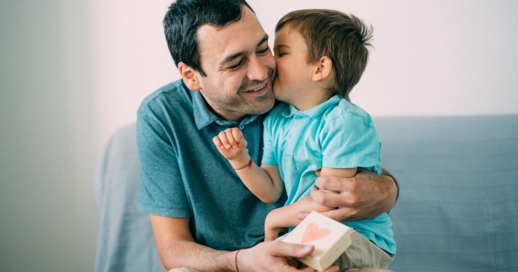 toddler fathers day gifts. Picture of a toddler kissing his dads cheek while his dad holds a small present.