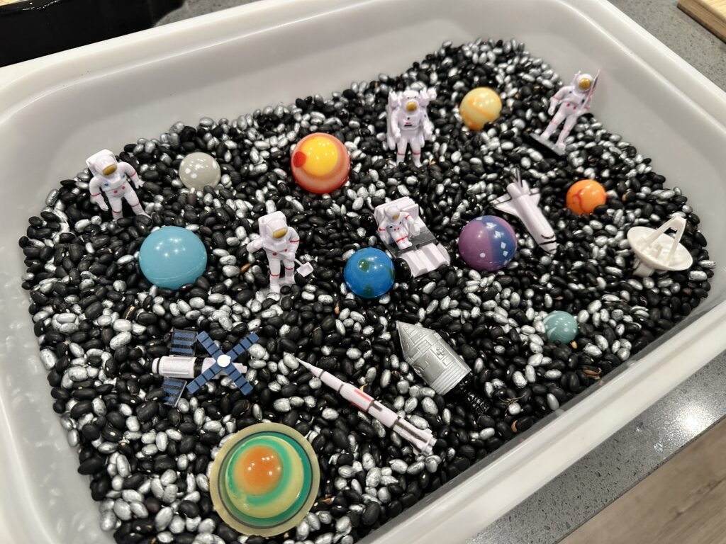 Finished space sensory bin with black and silver beans, glow in the dark planets, astronaut figurines, and a rocket ship